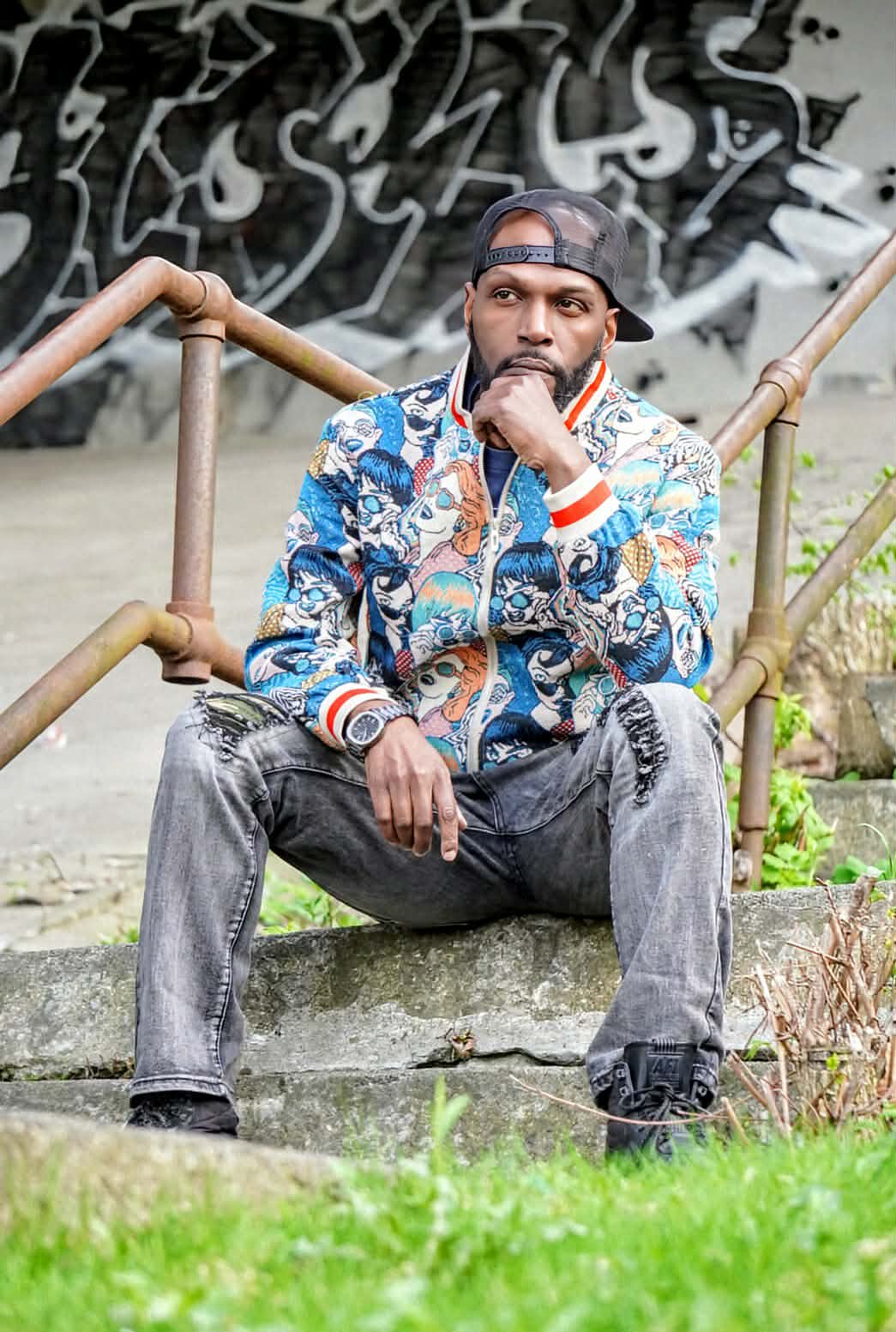 Wiz Hoffa seated on concrete steps between rusted pipe rails, hand on chin, backwards cap and faces-print bomber, black-and-white graffiti mural behind him