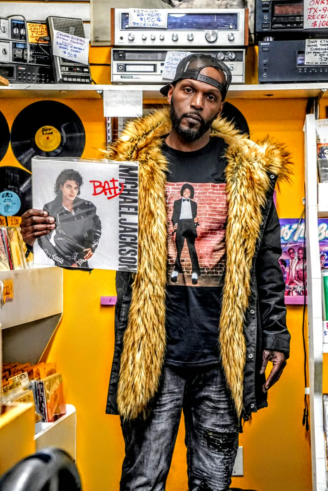 Wiz Hoffa in a record store holding Michael Jackson's "Bad" on vinyl, a wall of records and vintage receivers behind him