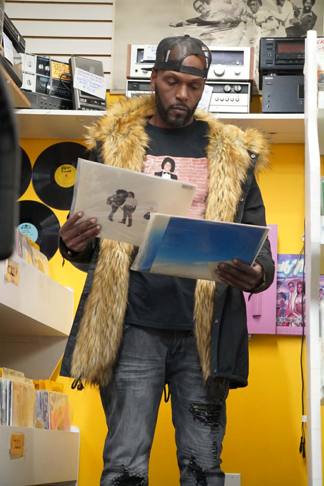 Wiz Hoffa standing in a record store examining two vinyl LPs, one in each hand, a wall of vinyl behind him