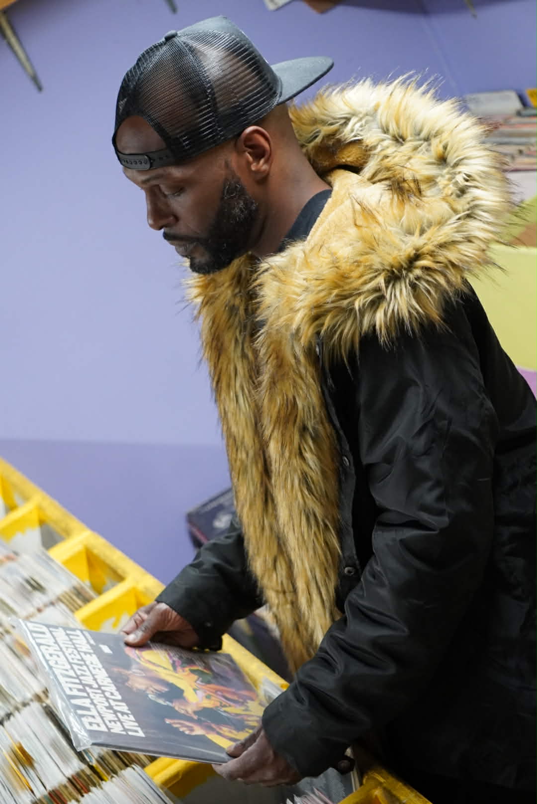Wiz Hoffa in profile at the record store crates, fur-collar hood up, pulling an album out of the stack