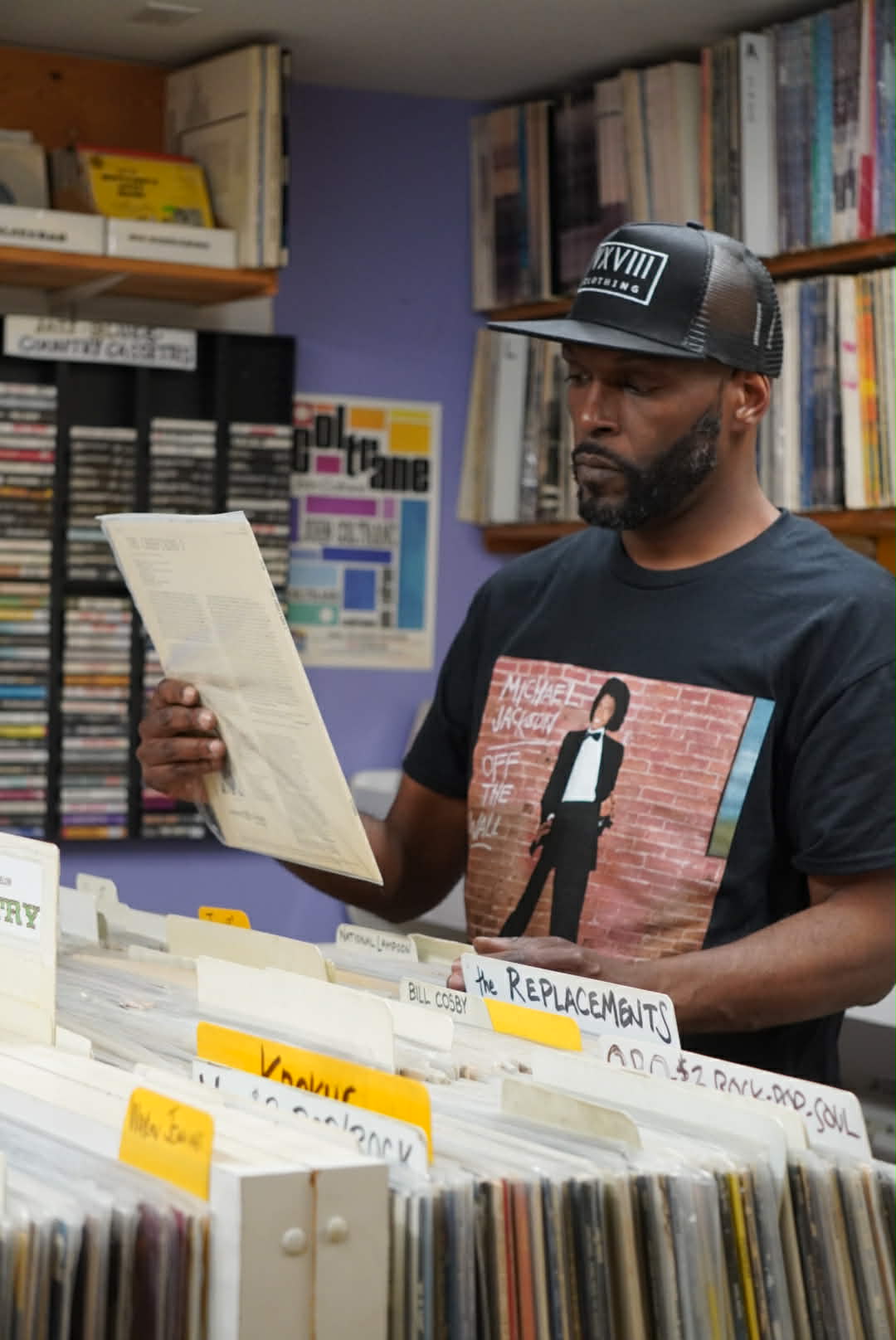 Wiz Hoffa in a Michael Jackson 'Off the Wall' tee and VXVIII cap, reading liner notes over record crates, cassette wall behind him