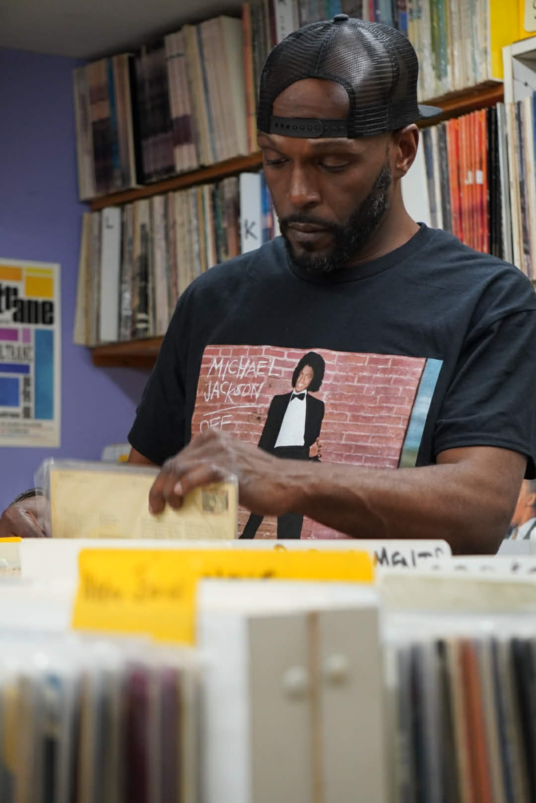 Closeup of Wiz Hoffa at the record crates in a backwards trucker cap, flipping a vinyl sleeve, shelves of records above him