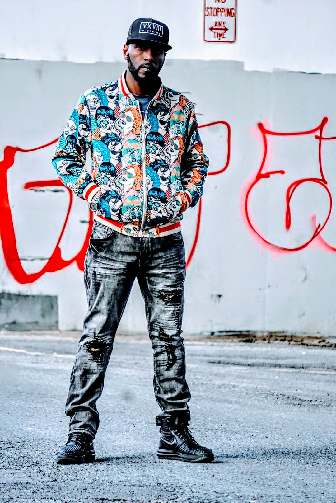 Wiz Hoffa standing in front of a white wall covered in huge red graffiti, hands in pockets, trucker cap and printed bomber jacket